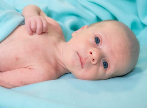 A newborn baby lies comfortably on a soft, aqua-colored blanket in a hospital room during the day. The baby gazes curiously at the camera, appearing calm and peaceful.