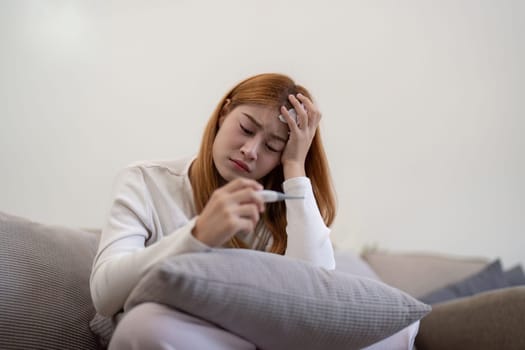 Young Asian woman feeling unwell and checking her temperature with a thermometer at home. Concept of illness, fever, and healthcare.