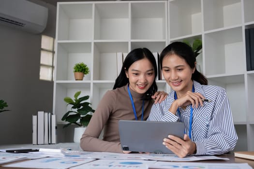 Two Asian businesswomen discussing work using a tablet in a modern office. Concept of teamwork and professional collaboration.