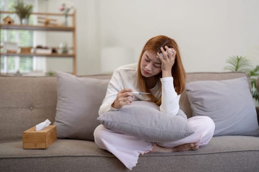 Young Asian woman feeling unwell and checking her temperature with a thermometer while sitting on the sofa at home. Concept of illness, fever, and healthcare.