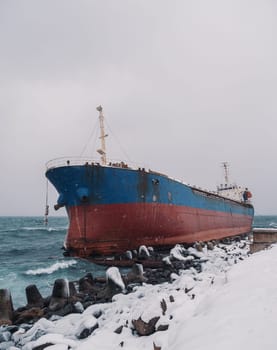 Cargo ship strands aground on a rocky shore after a storm, surrounded by snow and heavy waves.