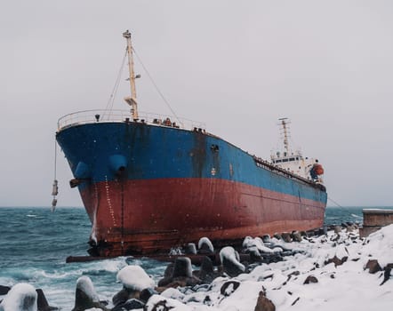 Cargo ship strands aground on a rocky shore after a storm, surrounded by snow and heavy waves.