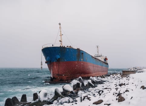 Cargo ship strands aground on a rocky shore after a storm, surrounded by snow and heavy waves.