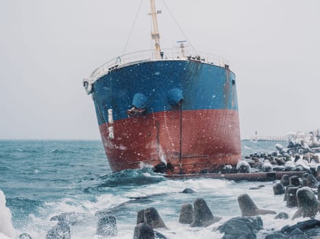 Cargo ship strands aground on a rocky shore after a storm, surrounded by snow and heavy waves.