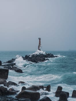 A solitary lighthouse stands firm on a rocky shore as waves crash against the rocks. The overcast sky and turbulent sea create a dramatic and moody atmosphere..