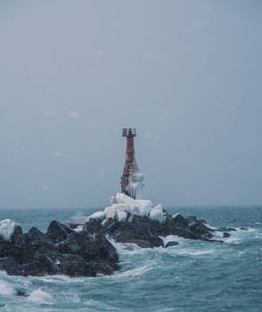 A solitary lighthouse stands firm on a rocky shore as waves crash against the rocks. The overcast sky and turbulent sea create a dramatic and moody atmosphere..