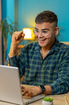 Indian man with laptop scream in delight triumph winner gesture celebrate success win money in lottery online casino game good news. Excited Hispanic guy sitting at home office workplace. Vertical