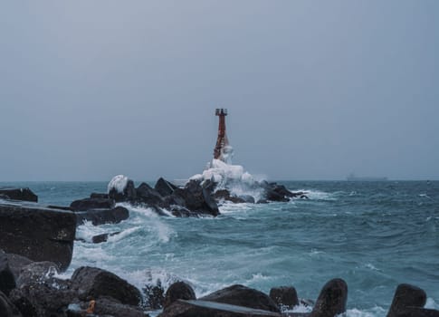 A solitary lighthouse stands firm on a rocky shore as waves crash against the rocks. The overcast sky and turbulent sea create a dramatic and moody atmosphere..