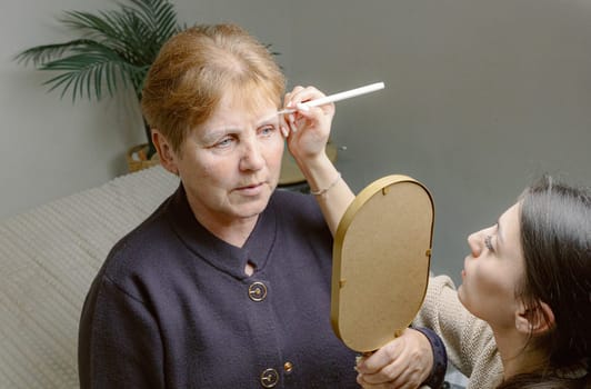 One young Caucasian elderly woman looks in the mirror, carefully watching as a girl cosmetologist draws a contour shape on her left eyebrow with a white marker, sitting in a home beauty salon on a cosmetology bed, close-up side view.