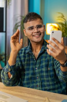 Happy Indian man blogger taking selfie on smartphone, communicating video call online with subscribers recording stories for social media. Hispanic guy sitting at home office table workplace. Vertical