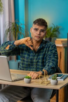 Dislike. Upset sad Indian business man at home office table showing thumbs down sign gesture, expressing discontent, disapproval dissatisfied bad work. Displeased Hispanic guy freelancer. Vertical