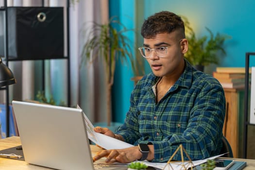 Indian freelancer man accountant working with financial graph documents calculate taxes for charts and reports at home office desk. Businessman guy working on laptop computer planning expenses budget