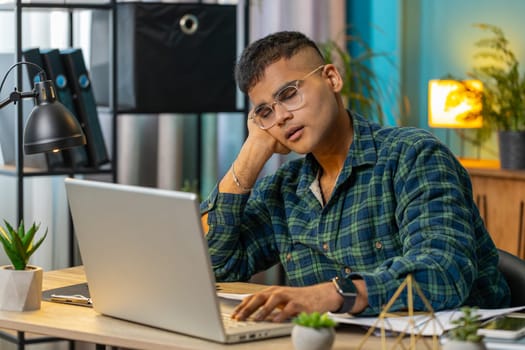 Bored sleepy Indian businessman worker working on laptop computer yawns, leaning on hand at home office desk. Exhausted tired man freelancer workaholic guy. Employment, occupation, workless. Lifestyle