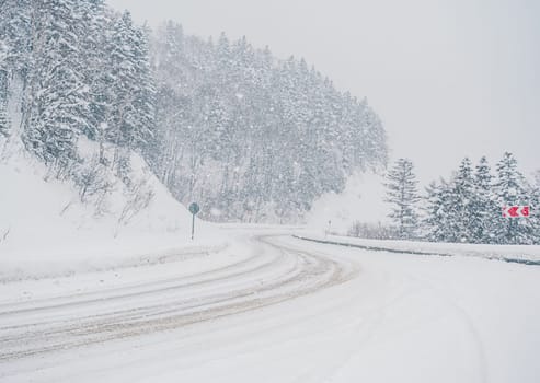 Snow is heavily falling on a winding mountain road lined with dense forest. The road appears slippery due to the accumulating snow.