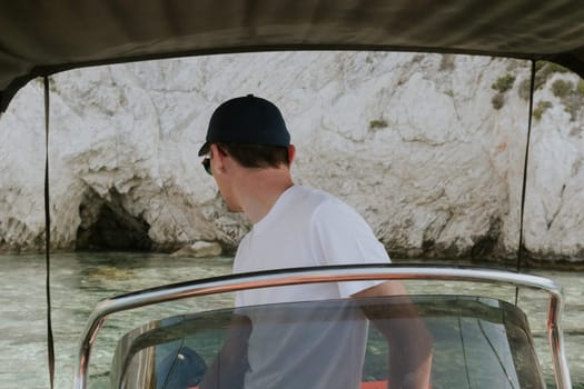 One young Caucasian guy in a cap and white T-shirt looks back and moored to a rocky beach, steering the helm on a sunny summer day, side view close-up.