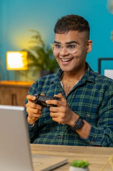 Excited Indian Arabian man playing video game on laptop spending leisure time at home office desk. Happy Hispanic freelancer guy playing video games with joystick controller on computer. Vertical