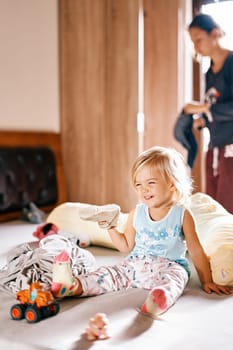 Little girl with a sneaker in her hand sits on a bed among toys. High quality photo