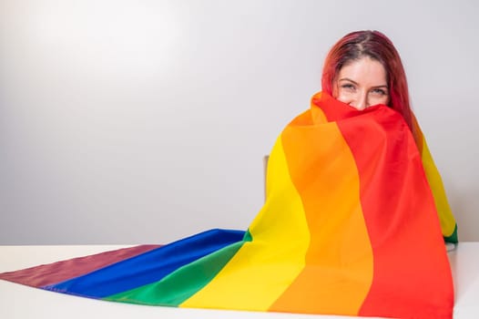 Young red-haired woman taking cover with lgbt flag on white background.