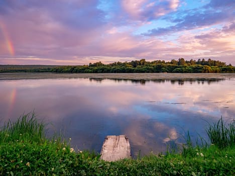 A serene countryside scene during sunset, showcasing a tranquil lake with a wooden dock in the foreground. A rainbow is reflected in the water, surrounded by a soft pink and purple sky.