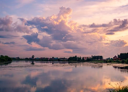 A tranquil river reflects the colorful sunset sky with clouds in a rural countryside setting. The serene scene is surrounded by lush greenery, creating a picturesque and peaceful environment.