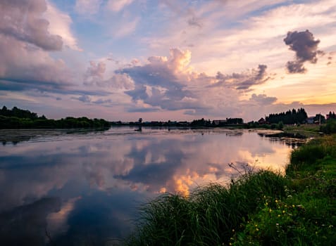 A tranquil river reflects the colorful sunset sky with clouds in a rural countryside setting. The serene scene is surrounded by lush greenery, creating a picturesque and peaceful environment.