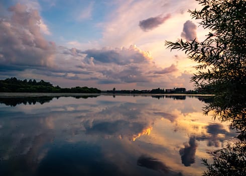 A tranquil river reflects the colorful sunset sky with clouds in a rural countryside setting. The serene scene is surrounded by lush greenery, creating a picturesque and peaceful environment.