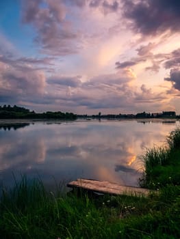 A serene countryside scene during sunset, showcasing a tranquil lake with a wooden dock in the foreground.