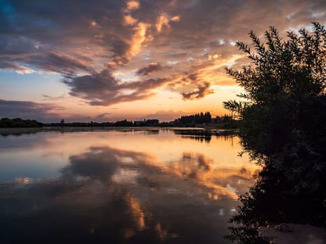 A tranquil river reflects the colorful sunset sky with clouds in a rural countryside setting. The serene scene is surrounded by lush greenery, creating a picturesque and peaceful environment.