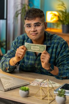 Indian man freelancer counting dollar cash insufficient amount of money. Financial crisis. Bankruptcy. Poverty and destitution. Hispanic male guy businessman with laptop sitting at home office table