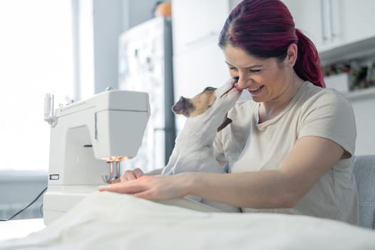 Caucasian woman sews while sitting in the kitchen. Dog Jack Russell Terrier sits on the lap of the owner. Home hobby