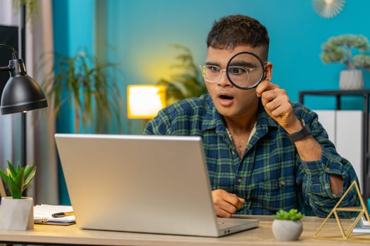 Surprised investigator researcher Indian businessman working at home office holding magnifying glass looking at laptop screen with wow expression, searching, analyzing. Amazed shocked freelancer guy