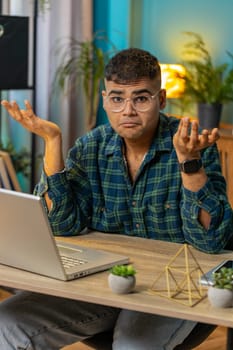 I don't know what to say. Confused Indian man feeling embarrassed about ambiguous question, having doubts, no idea, being clueless and uncertain. Hispanic guy at home office table workplace. Vertical