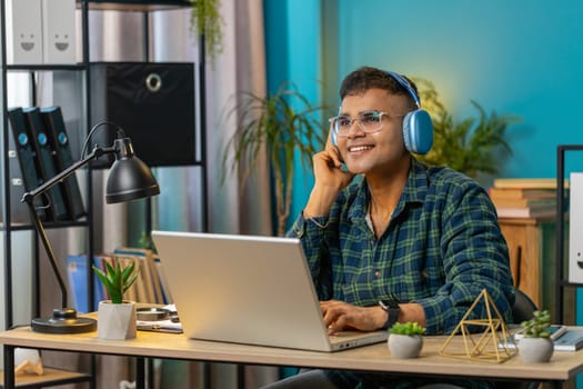 Happy overjoyed Indian man sits at home office desk listening to energetic disco music through wireless headphones. Arabian freelancer guy relaxing taking a break with laptop on table. Lifestyles