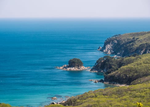 A stunning coastal landscape showcases rugged cliffs, lush green vegetation, and a clear blue sea on a bright sunny day.