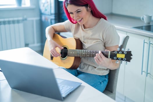 A woman sits in the kitchen during a remote acoustic guitar lesson. A girl learns to play the guitar and watches educational videos on a laptop.