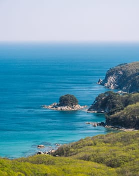 A stunning coastal landscape showcases rugged cliffs, lush green vegetation, and a clear blue sea on a bright sunny day.