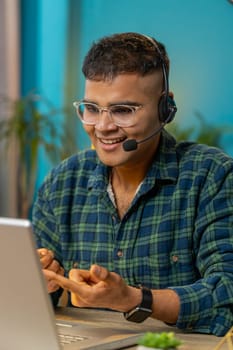 Indian business man wearing headset freelance worker call center or support service operator helpline talking with client or colleague communication support. Guy sitting at home office desk. Vertical