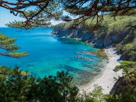 A picturesque, calm cove bordered by a rocky coastline and lush, green forest. The turquoise blue waters glisten under the midday sun, creating a serene and inviting atmosphere. Pine trees frame the scene, adding to the natural beauty.