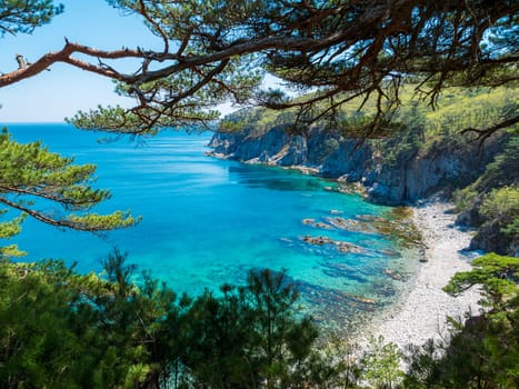 A picturesque, calm cove bordered by a rocky coastline and lush, green forest. The turquoise blue waters glisten under the midday sun, creating a serene and inviting atmosphere. Pine trees frame the scene, adding to the natural beauty.