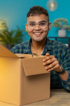 Happy Indian man unpacking delivery parcel. Smiling satisfied Hindu guy shopper online shop customer opening cardboard box receiving purchase gift by fast postal shipping at home office desk. Vertical