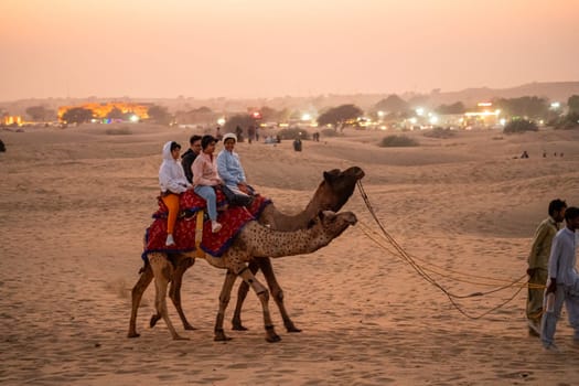 Jaisalmer, Rajasthan, India - 25th Dec 2023: Muslim man in traditional clothes leading camel with tourists across thar desert in Sam Jaisalmer Rajasthan at sunset showing this popular activity