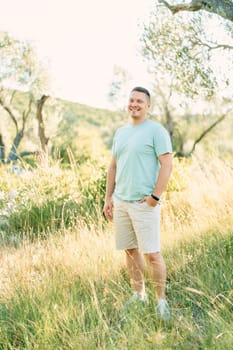 Smiling guy with hand in shorts pocket standing in sunny park. High quality photo