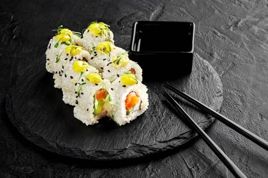 Sesame coated sushi rolls with avocado and fresh salmon topped with drop of mayo and microgreens traditionally served with soy sauce on black slate board. Japanese cuisine