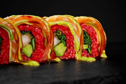 Closeup of vibrant red rice sushi rolls with tempura shrimp and avocado, topped with salmon slice and spicy sauces, presented on black slate board. Japanese fusion cuisine