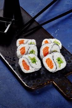Set of yin and yang shaped futomaki rolls filled with salmon and crisp cucumber served on textured plate with chopsticks and soy sauce against blue background. Japanese style snack