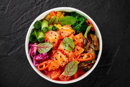 Colorful poke bowl with salmon, grilled zucchini, sliced fresh vegetables, hiyashi wakame and greens sprinkled with sesame in cardboard cup on dark surface. Hawaiian style meal. Takeaway food concept