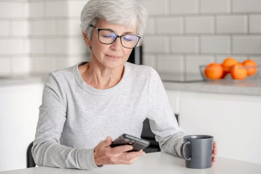 Grey-Haired Attractive Woman Searches For Information On Phone At Home At Table