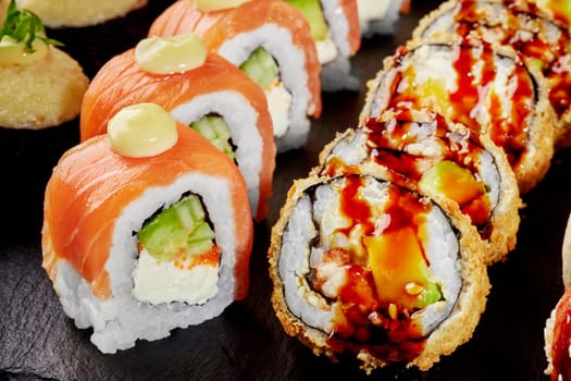 Closeup of delicious crispy tempura roll filled with eel drizzled with tangy unagi sauce and uramaki with fresh salmon and tobiko seasoned with spicy mayo, served on slate board. Japanese cuisine