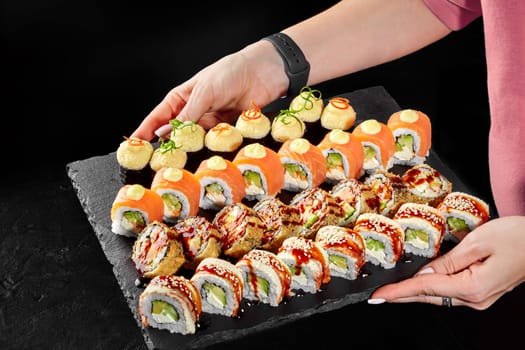 Female hands holding serving slate board brimming with sushi variety, featuring eel and salmon wrapped uramaki, crispy tempura roll drizzled with tangy unagi sauce, and baked norimaki with cheese caps