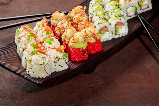 Enticing set of sushi rolls with salmon, shrimp, tobiko and sesame garnished with greens and spicy sauce, arranged on dark wooden serving tray. Stylish presentation of popular Japanese dish